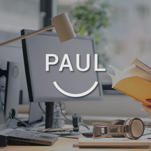 Schreibtisch mit einem Monitor und einer Schreibtischlampe. Mittig im Bild wird das Mein Paul Logo dargestellt.