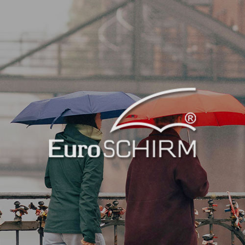 Zwei Frauen laufen bei Regen auf einer Brücke mit jeweils einem Regenschirm in der Hand. Mittig im Bild wird das Euroschirm Logo dargestellt.