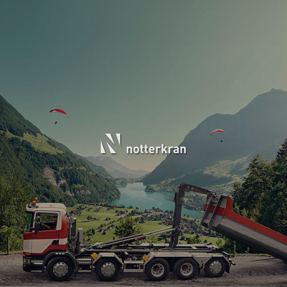 Lastwagen fährt auf einer Strasse und im Hintergrund sind Berge. Mittig im Bild wird das Notterkran Logo dargestellt.