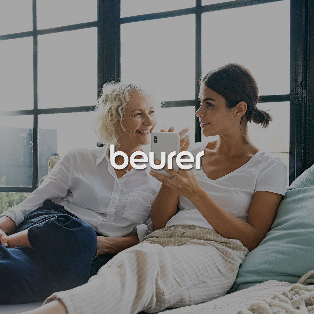 Zwei lächelnde Frauen sitzen auf dem Sofa und schauen auf ein Handy. Mittig im Bild wird das beurer Logo dargestellt.