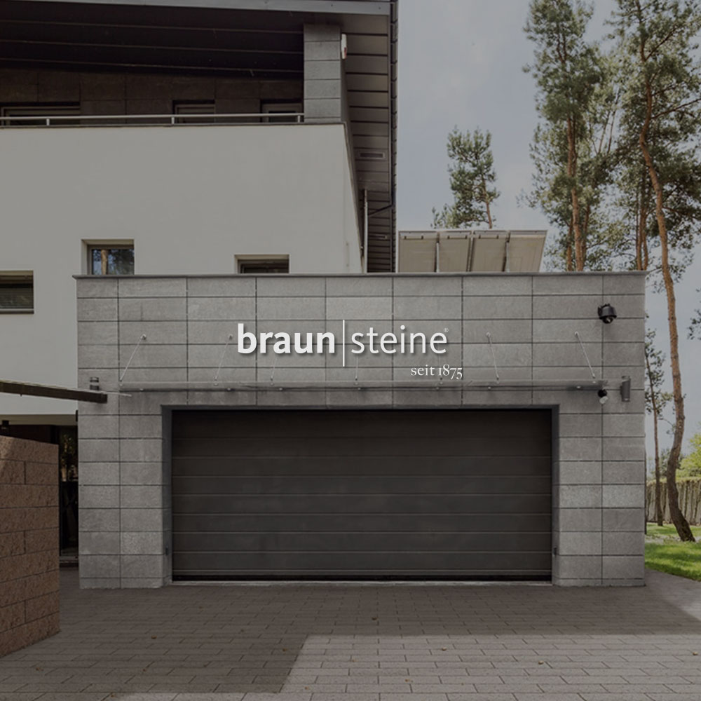 Haus mit einer Garage die aus grauen Steinen besteht. Mittig im Bild wird das Braun-Steine Logo dargestellt.