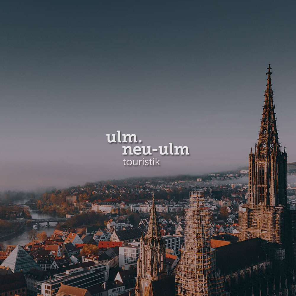 Blick von oben auf die Stadt Ulm und das Ulmer Münster. Mittig im Bild wird das Ulm / Neu-Ulm Touristik Logo dargestellt.